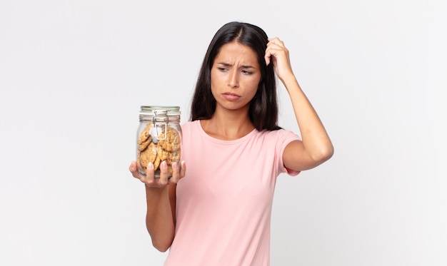 Jonge Spaanse vrouw die zich verward en verward voelt, haar hoofd krabt en een glazen fles met koekjes vasthoudt