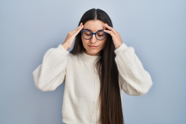 Jonge spaanse vrouw draagt een casual trui over blauwe achtergrond met de hand op het hoofd voor pijn in het hoofd omdat stress lijdt aan migraine