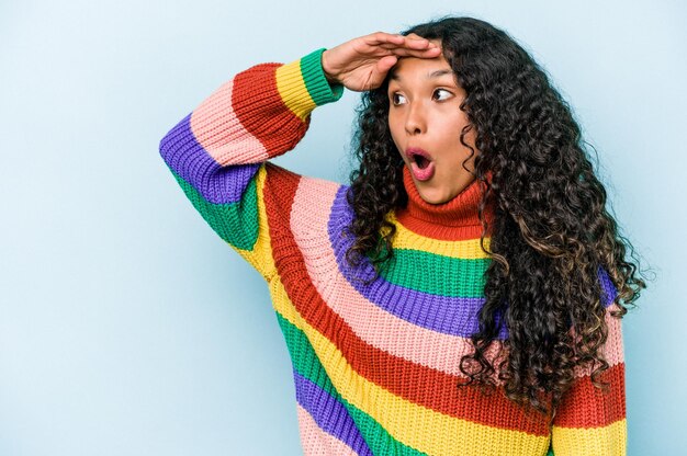 Jonge Spaanse vrouw geïsoleerd op een blauwe achtergrond die ver weg kijkt en hand op het voorhoofd houdt