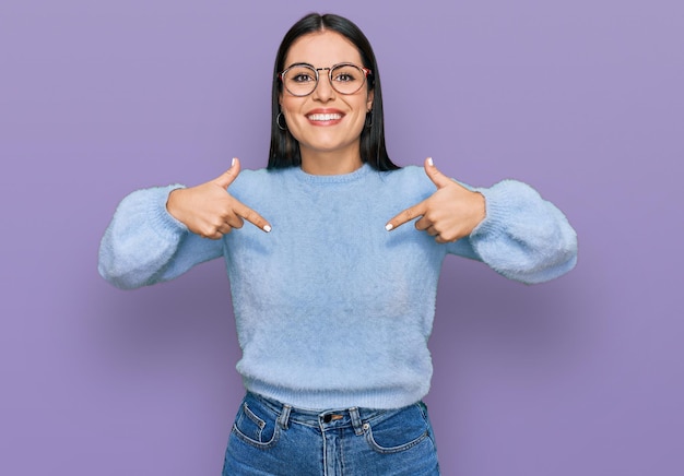 Jonge Spaanse vrouw, gekleed in casual kleding en een bril, zelfverzekerd met een glimlach op het gezicht, zichzelf wijzend met vingers, trots en gelukkig