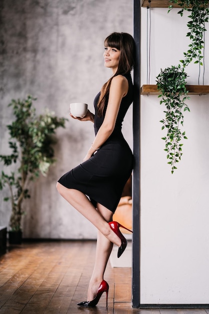 Jonge spaanse vrouw gekleed in zwarte elegante jurk thuis leunt tegen muur met plant op de planken