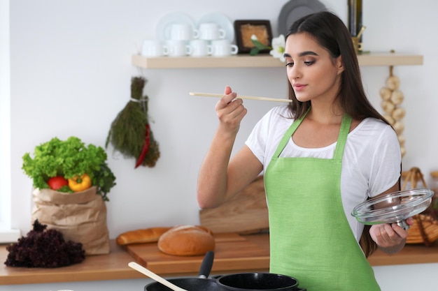 Jonge Spaanse vrouw in een groene schort die in de keuken kookt terwijl ze op een houten lepel blaast Huisvrouw vond een nieuw recept voor haar soep