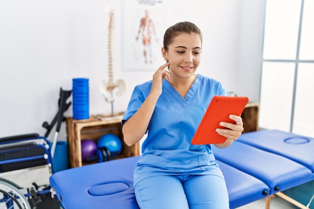 Jonge spaanse vrouw in fysiotherapie-uniform met videogesprek in sportcentrum