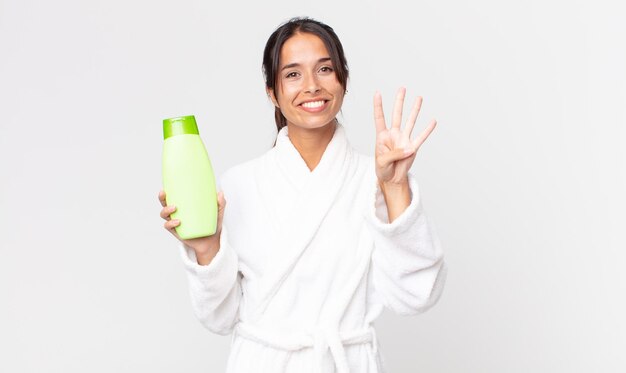 Jonge Spaanse vrouw lacht en ziet er vriendelijk uit en toont nummer vier in een badjas en met een shampoo