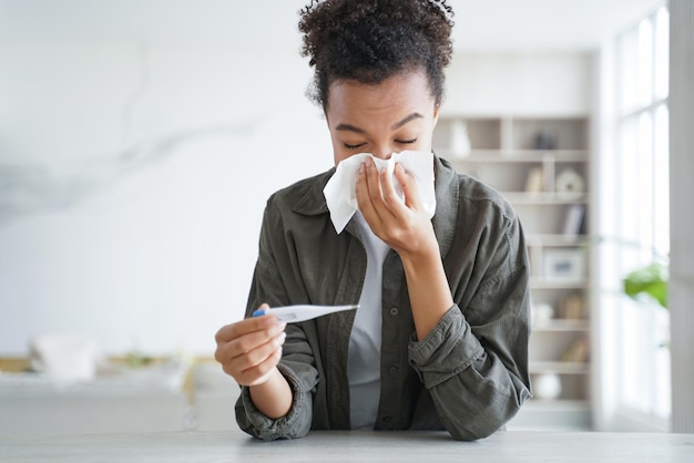 Jonge Spaanse vrouw meet de temperatuur met een thermometer die niest en een loopneus blaast