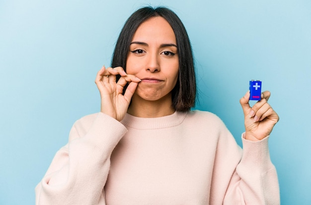 Jonge Spaanse vrouw met batterijen geïsoleerd op een blauwe achtergrond met vingers op de lippen die een geheim houden