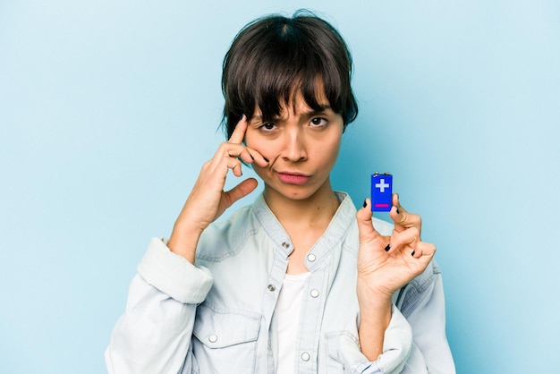 Jonge Spaanse vrouw met een batterij geïsoleerd op een blauwe achtergrond wijzende tempel met vinger denken gericht op een taak