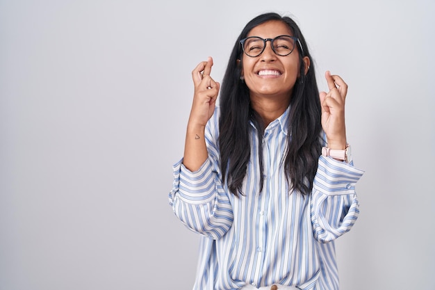 Jonge Spaanse vrouw met een bril gebaren vinger gekruist lachend met hoop en gesloten ogen geluk en bijgelovig concept
