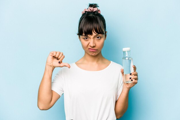 Jonge Spaanse vrouw met een fles water geïsoleerd op een blauwe achtergrond met een afkeer gebaar duimen omlaag meningsverschil concept