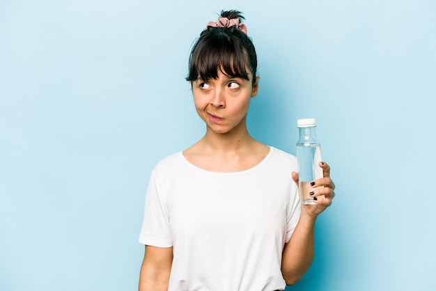 Jonge Spaanse vrouw met een fles water geïsoleerd op een blauwe achtergrond verward voelt zich twijfelachtig en onzeker