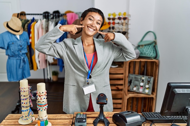 Jonge spaanse vrouw met kort haar werkend als manager bij kleinhandelsboutique glimlachend vrolijk tonend en wijzend met vingerstanden en mond tandheelkundig gezondheidsconcept