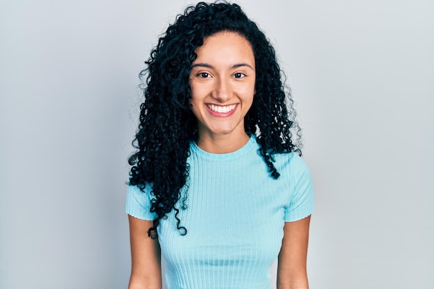 Jonge Spaanse vrouw met krullend haar die een casual blauw t-shirt draagt met een gelukkige en koele glimlach op haar gezicht.