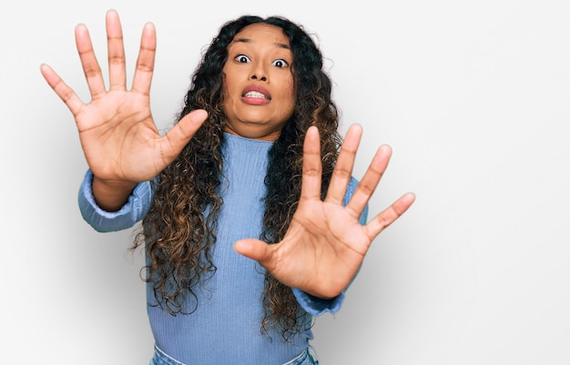 Jonge spaanse vrouw met krullend haar, gekleed in vrijetijdskleding, bang en doodsbang van angstuitdrukking, stop gebaar met handen schreeuwend in shock paniek concept