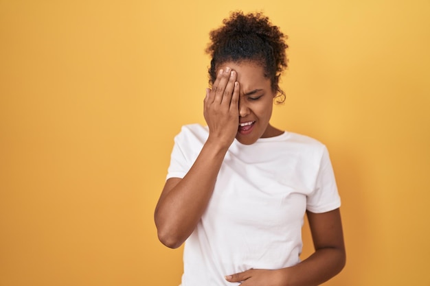 Jonge spaanse vrouw met krullend haar staande over gele achtergrond geeuwen moe bedekkend half gezicht oog en mond met hand gezicht doet pijn van pijn