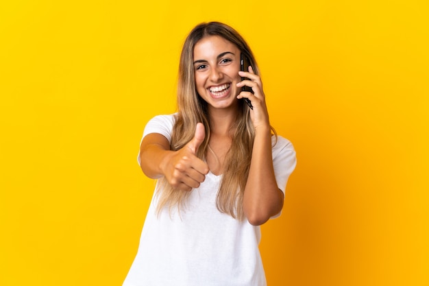 Jonge Spaanse vrouw over geïsoleerd geel die een gesprek met mobiel houdt terwijl het doen van duimen omhoog