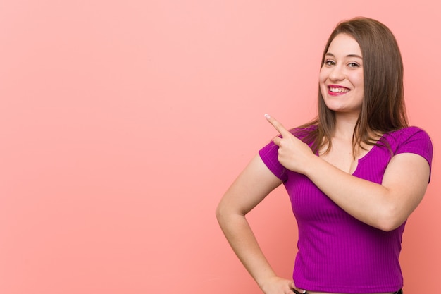 Jonge Spaanse vrouw tegen een roze en muur die opzij glimlacht richt iets op lege ruimte toont.