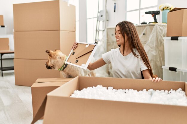 Jonge spaanse vrouw unboxing pakket zittend op de vloer met hond in nieuw huis