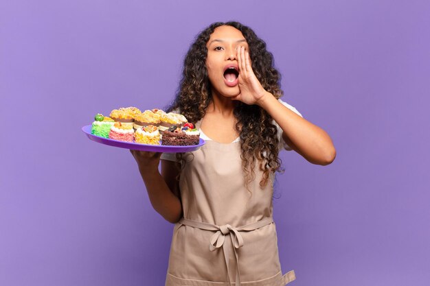 Foto jonge spaanse vrouw voelt zich gelukkig opgewonden en positief en geeft een grote schreeuw met handen naast de mond en roept het concept van kookcakes op