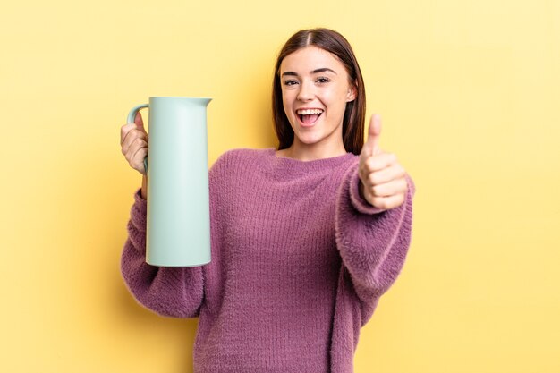 Jonge spaanse vrouw voelt zich trots, positief glimlachend met duimen omhoog. koffie thermos concept