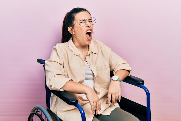 Jonge spaanse vrouw zittend op rolstoel boos en boos schreeuwend gefrustreerd en woedend, schreeuwend van woede. woede en agressief concept.