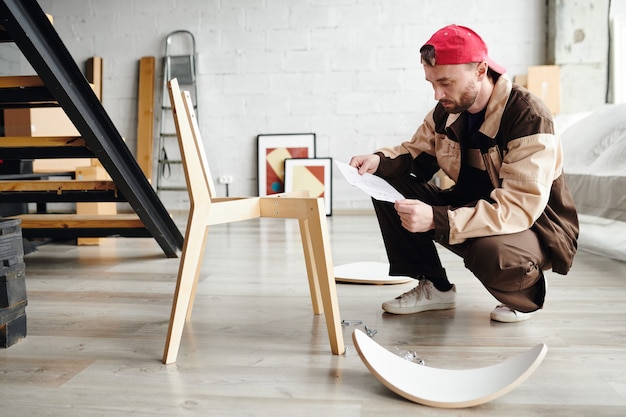 Jonge specialist in meubelassemblage die op kraakpanden staat en instructies op papier doorneemt terwijl hij een nieuwe houten stoel in elkaar zet