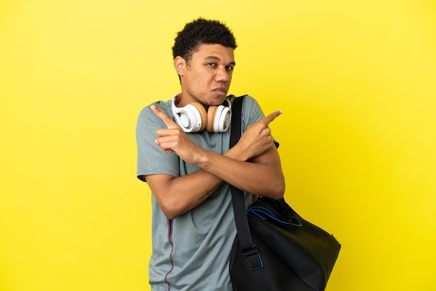Jonge sport Afro-Amerikaanse man met sporttas geïsoleerd op gele achtergrond wijzend naar de zijtakken die twijfels hebben