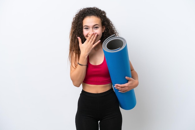 Jonge sport Arabische vrouw die naar yogalessen gaat terwijl ze een mat vasthoudt op een witte achtergrond, gelukkig en glimlachend die mond bedekt met hand