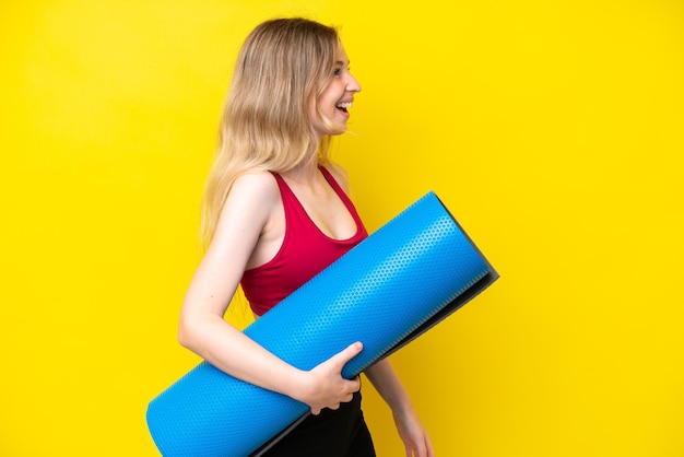 Jonge sport blanke vrouw die naar yogalessen gaat terwijl ze een mat vasthoudt geïsoleerd op gele achtergrond lachend in laterale positie