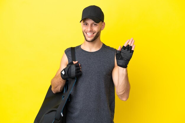 Jonge sport blonde man met sporttas geïsoleerd op gele achtergrond saluerend met de hand met gelukkige uitdrukking