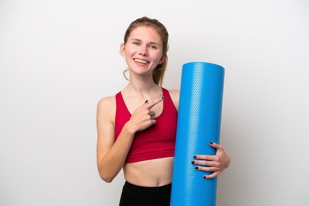 Jonge sport Engelse vrouw die naar yogalessen gaat terwijl ze een mat vasthoudt op een witte achtergrond die naar de zijkant wijst om een product te presenteren