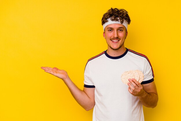 Jonge sport Kaukasische man die een rijstwafel eet die op gele achtergrond wordt geïsoleerd die een exemplaarruimte op een palm toont en een andere hand op taille houdt.