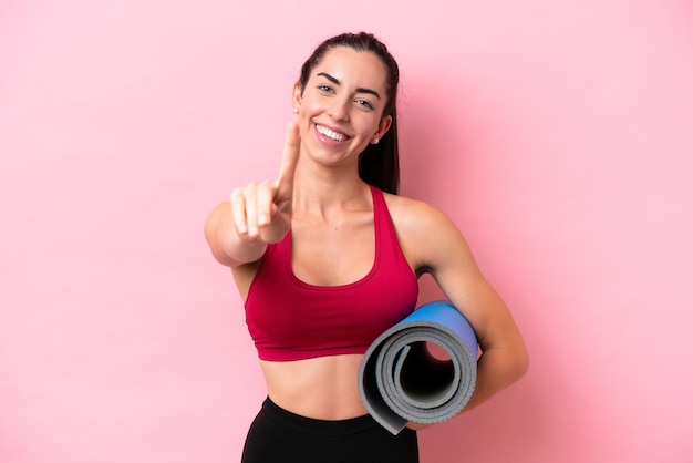 Jonge sport-kaukasische vrouw die naar yogalessen gaat terwijl ze een mat vasthoudt op een roze achtergrond die een vinger laat zien en optilt