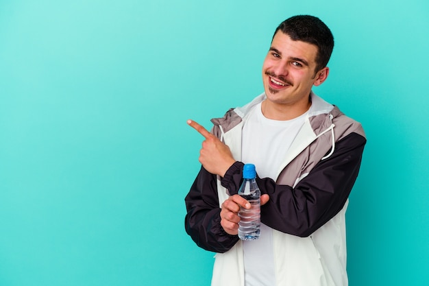 Jonge sportieve blanke man drinkwater geïsoleerd op blauwe muur glimlachend en opzij wijzend, iets laten zien op lege ruimte.