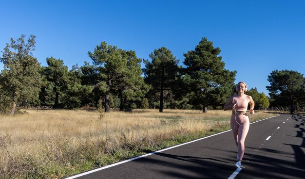 Jonge sportieve vrouw die op een verharde weg loopt en naar muziek luistert Glimlachend atleet meisje dat op een weg loopt en naar muziek luistert Concept van gezond leven en vrouw die op de weg loopt