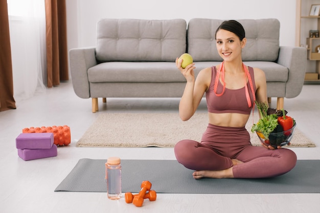 Jonge sportieve vrouw die op yogamat zit en een verscheidenheid aan groenten en appelconcept vasthoudt van gewichtsverlies en gezond eten