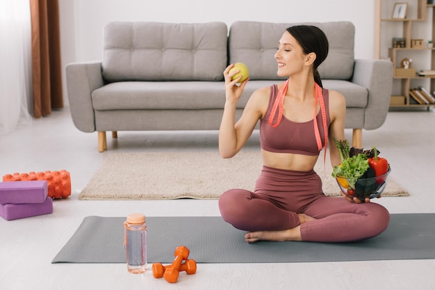 Jonge sportieve vrouw die op yogamat zit en een verscheidenheid aan groenten en appelconcept vasthoudt van gewichtsverlies en gezond eten