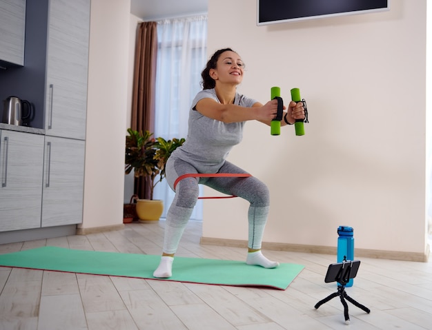 Jonge sportieve vrouw dumbbell squats met elastische fitness band uitvoeren tijdens het kijken naar training tutorials. Thuis trainen