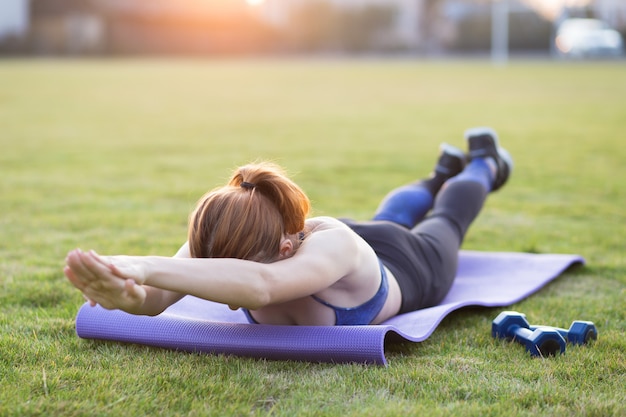 Jonge sportieve vrouw in sportkleren die op gebied bij zonsopgang opleiden. Meisje dat zich in plankpositie bevindt over gras in een stadspark.