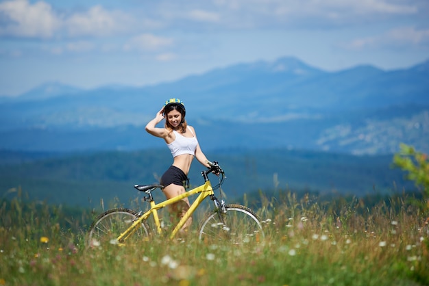 Jonge sportieve vrouwelijke fietser die zich met gele bergfiets bevinden op een gras, op de zomerdag. Bergen en blauwe lucht op de achtergrond. Buitensport lifestyle activiteit.