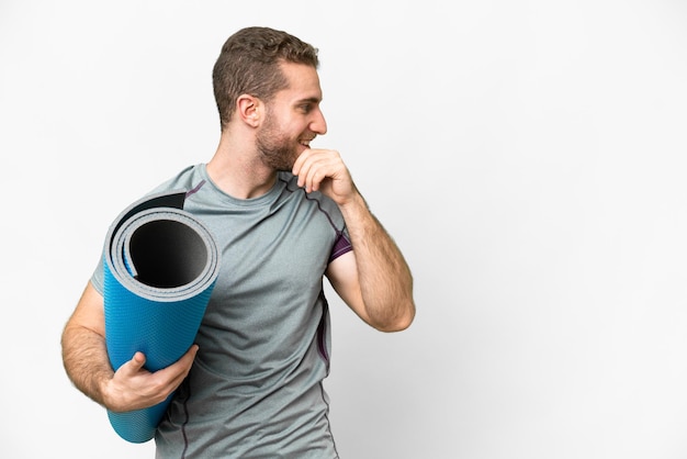 Jonge sportman die naar yogalessen gaat terwijl hij een mat over een geïsoleerde witte achtergrond houdt, een idee denkt en naar de zijkant kijkt