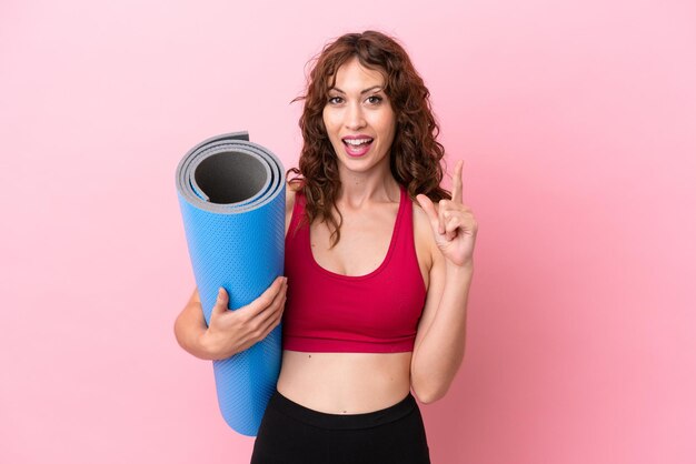 Jonge sportvrouw die naar yogalessen gaat terwijl ze een mat vasthoudt die op een roze achtergrond is geïsoleerd en denkt aan een idee met de vinger omhoog