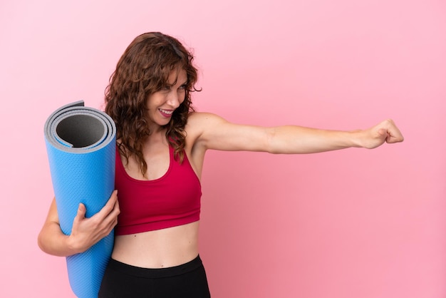 Jonge sportvrouw die naar yogalessen gaat terwijl ze een mat vasthoudt die op een roze achtergrond wordt geïsoleerd en een duim omhoog gebaar geeft