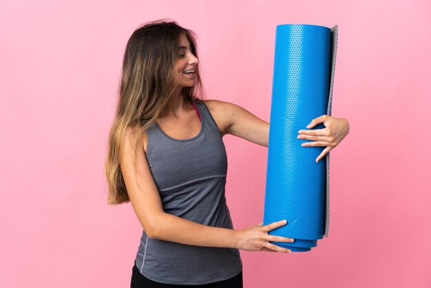 Jonge sportvrouw die naar yogalessen gaat terwijl ze een mat vasthoudt die op roze muur met gelukkige uitdrukking wordt geïsoleerd