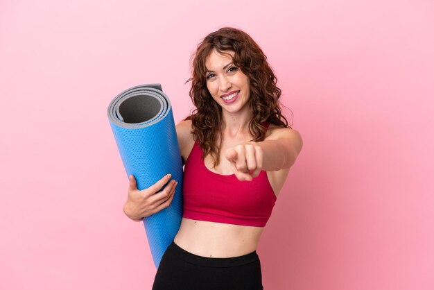 Jonge sportvrouw die naar yogalessen gaat terwijl ze een mat vasthoudt op een roze achtergrond die naar voren wijst met een gelukkige uitdrukking