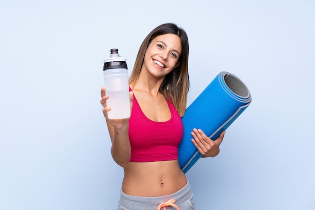 Jonge sportvrouw over geïsoleerd blauw met de fles van het sportenwater en met een mat