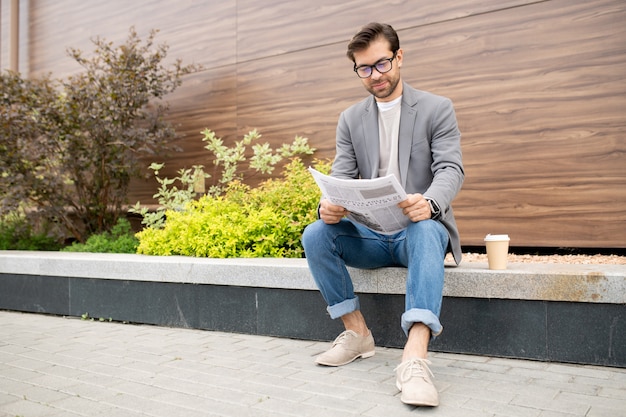 Jonge stedelijke zakenman in spijkerbroek en jas zitten in een stedelijke omgeving en de krant lezen met het laatste nieuws