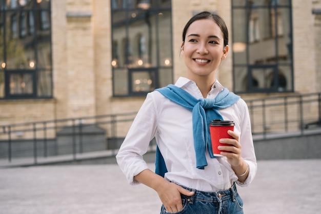 Jonge stijlvolle aziatische vrouw met kopje koffie, staande op stedelijke straat, taxi wachten