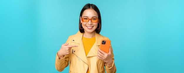 Jonge stijlvolle Aziatische vrouwelijke model in trendy zonnebril lente outfit met mobiele telefoon wijzend op smartphone en glimlachend staande over blauwe achtergrond