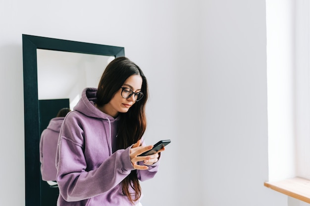 Jonge stijlvolle blanke vrouw met behulp van mobiele telefoon in de buurt van grote spiegel, zittend op een zwarte stoel in witte kamer in de buurt van raam.