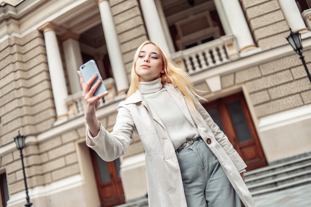 Jonge stijlvolle blonde vrouw maakt selfie op smartphone in de stad